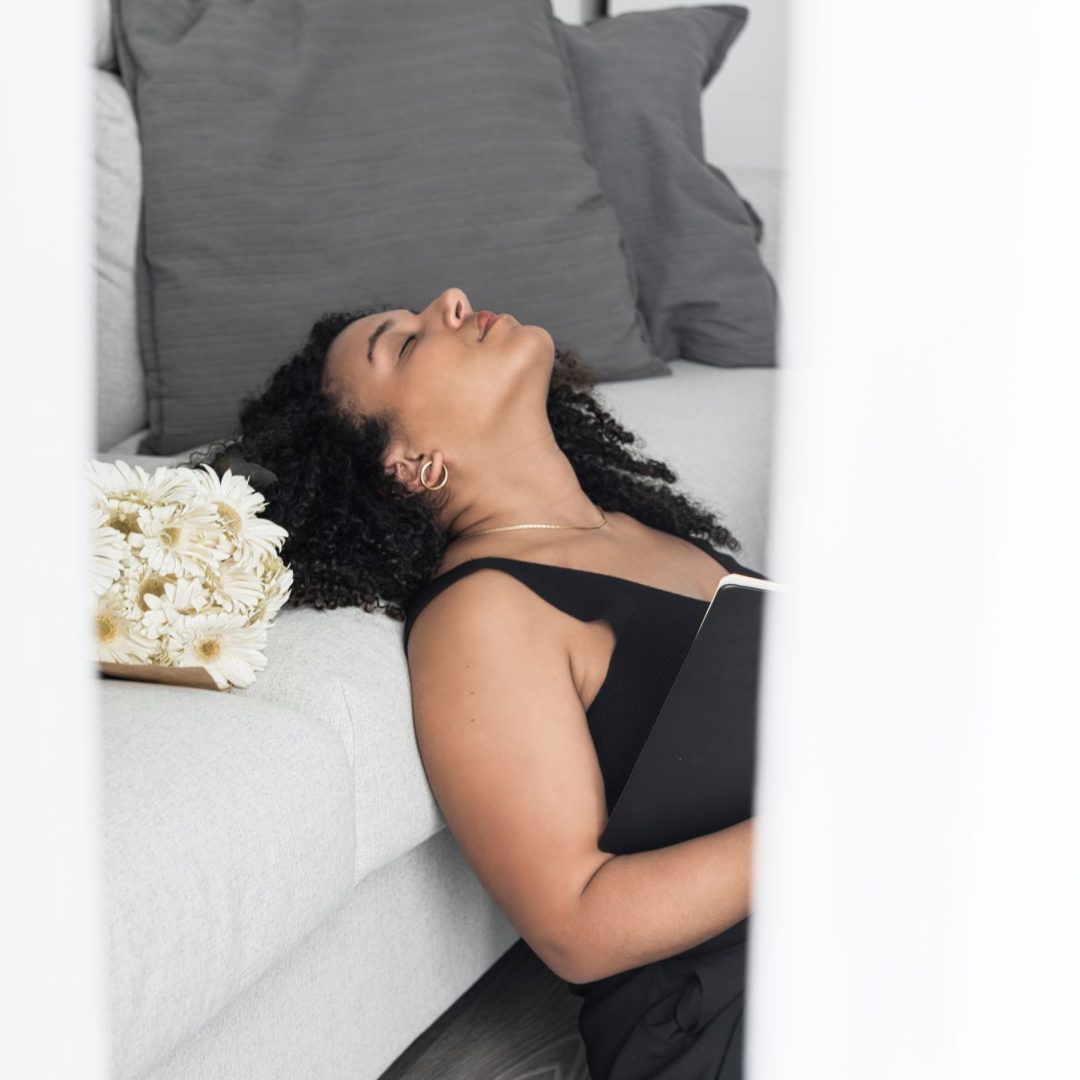 Mulher com a cabeça encostada no sofá e olhos fechados, um ramo de flores encontra-se ao lado da sua cabeça.